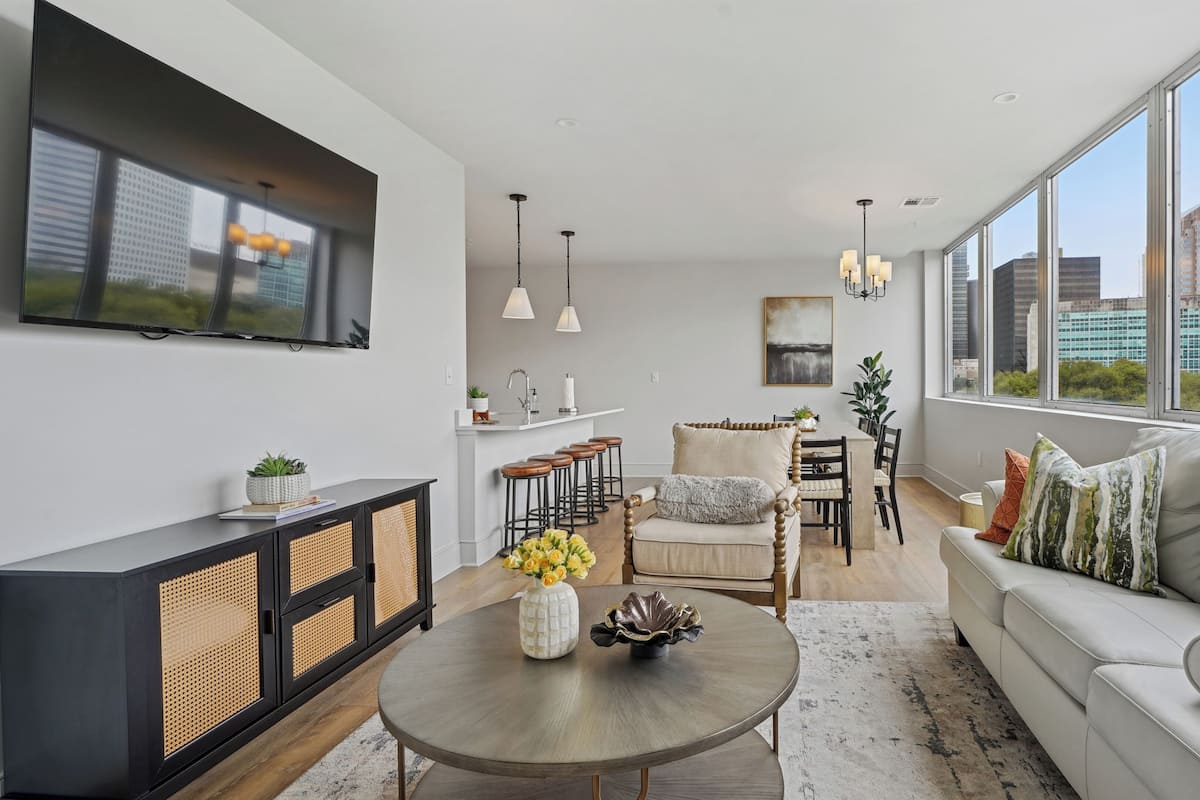 Un salon moderne avec un canapé, une table basse et un coin repas. Un téléviseur est fixé au mur, et de grandes fenêtres offrent une vue sur la ville.