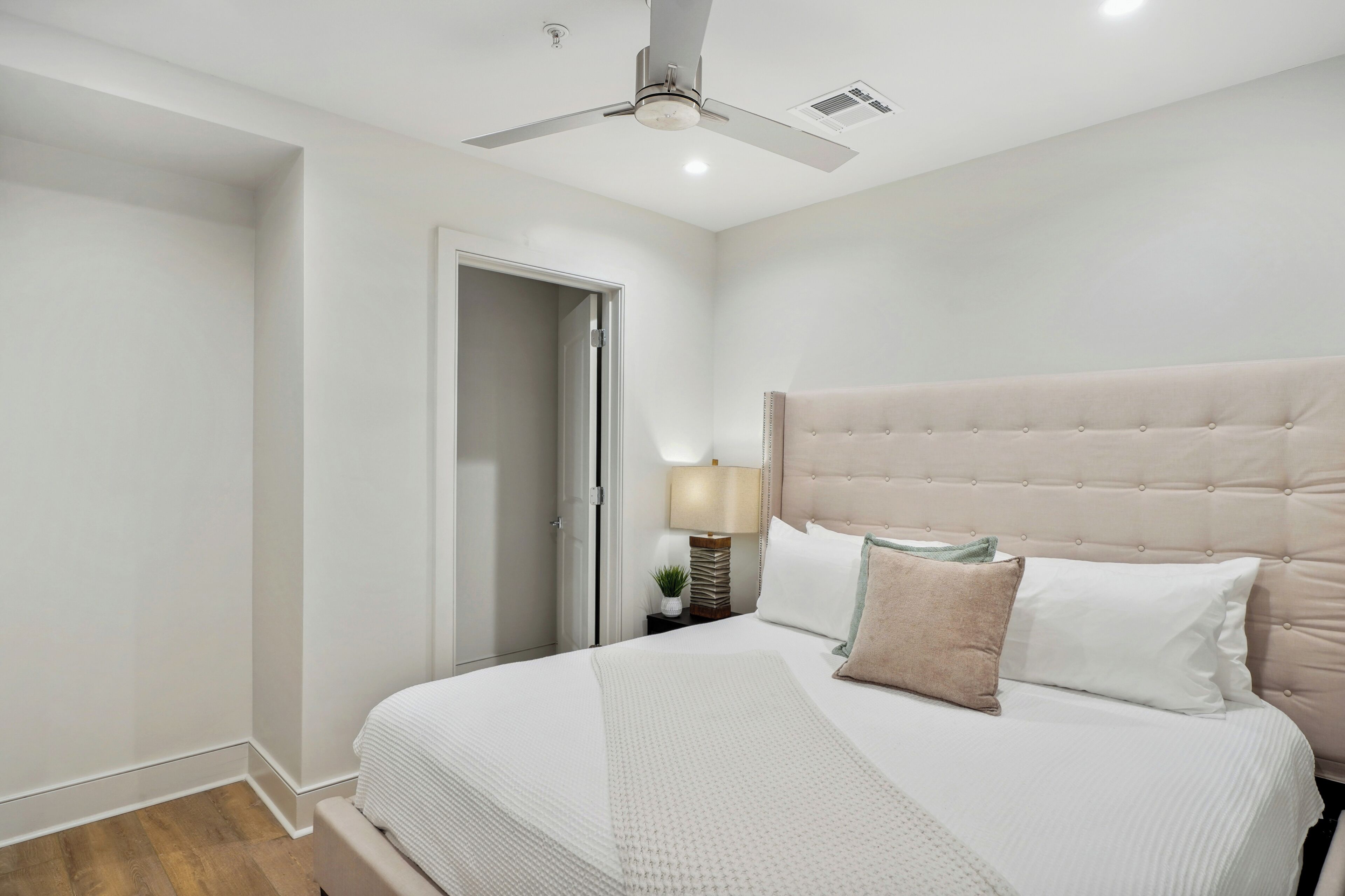 Ein Schlafzimmer mit einem großen Bett, einem Deckenventilator, einem Nachttisch mit Lampe und einer Tür zu einem weiteren Raum.