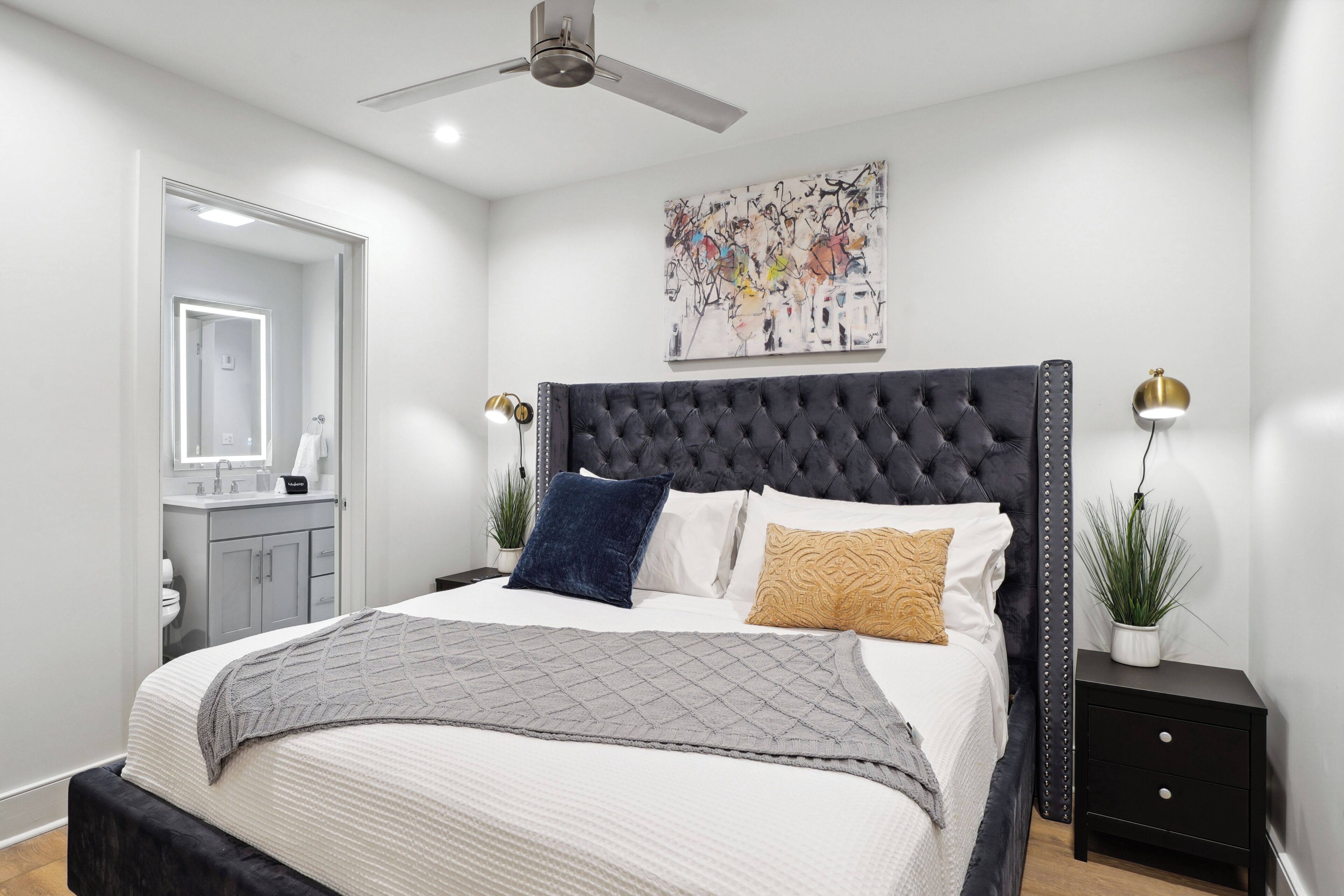 Una camera da letto moderna con testiera imbottita, un letto grande con copripiumino grigio, un comodino con lampada e un'opera d'arte appesa al muro.