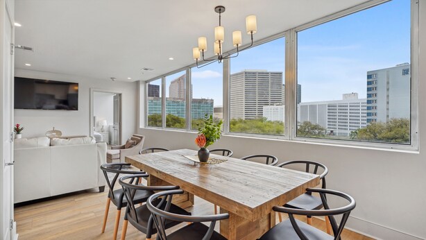 A modern dining area with a wooden table and chairs, a vase with flowers, and large windows offering a city view.