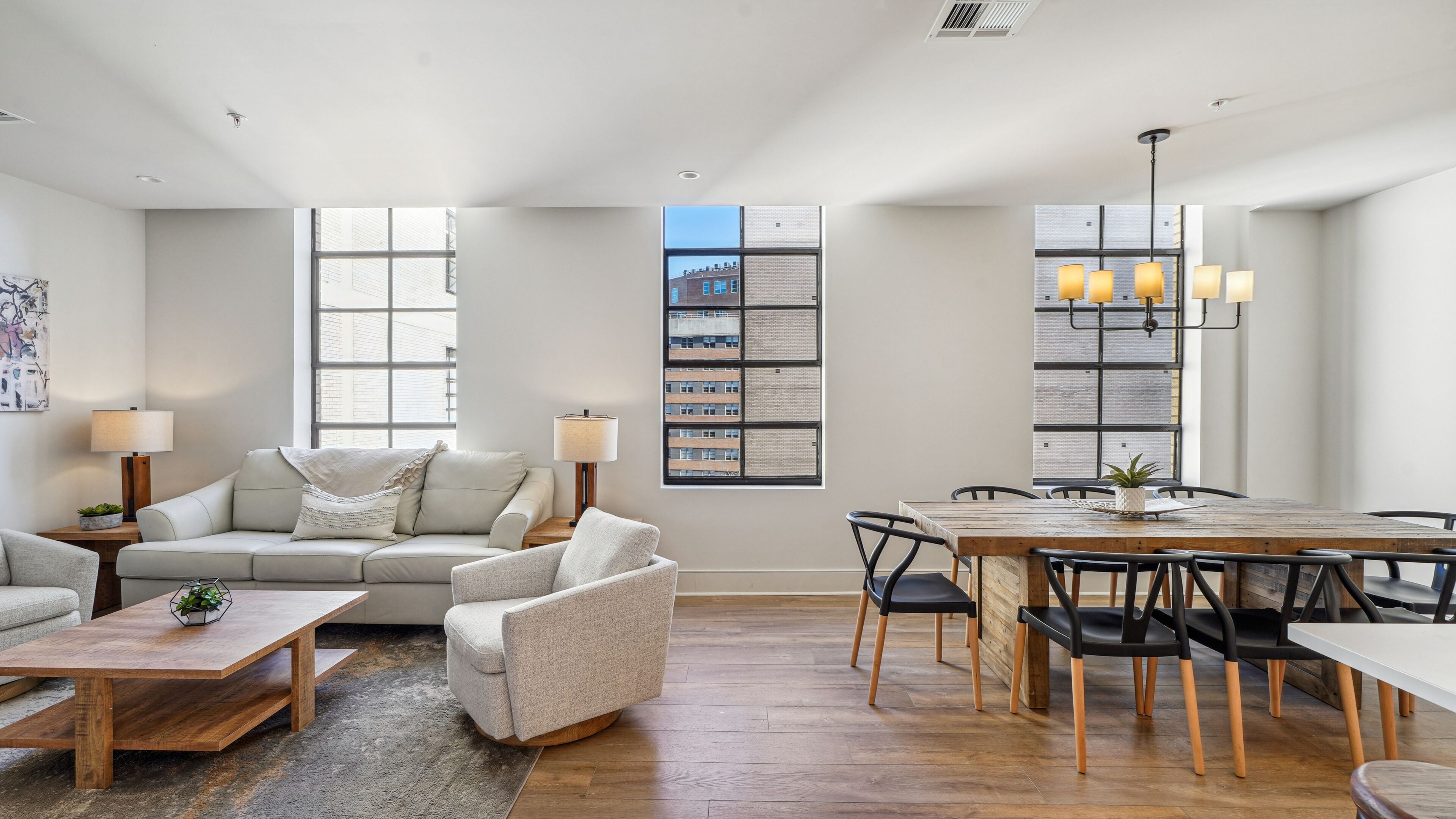 A spacious living room with a sofa, armchair, and dining table set. There are large windows with city views, a bookshelf, and a lamp on the side table.
