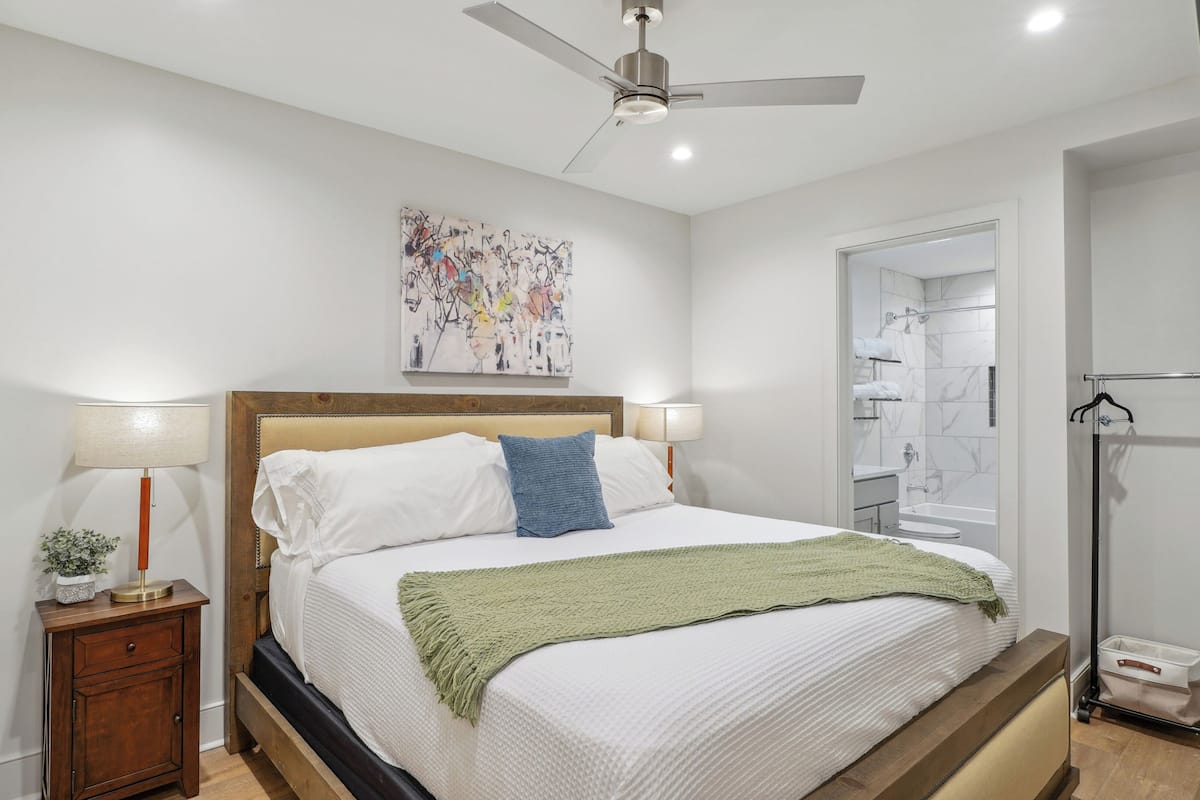 Une chambre à coucher avec un lit, des lampes de chevet, un ventilateur de plafond, un tableau et une salle de bain visible à travers une porte ouverte.