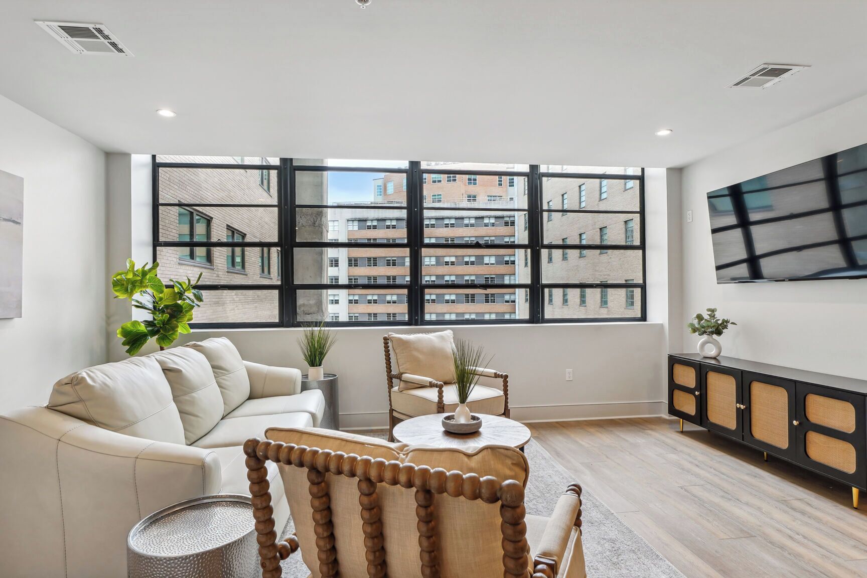 A modern living room with a sofa, armchair, and a coffee table. There is a large window with a view of buildings.