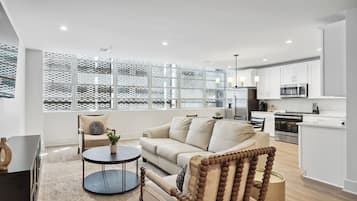 A modern living room with a sofa, a round coffee table, and a chair. There is a kitchen area with white cabinets and a microwave.