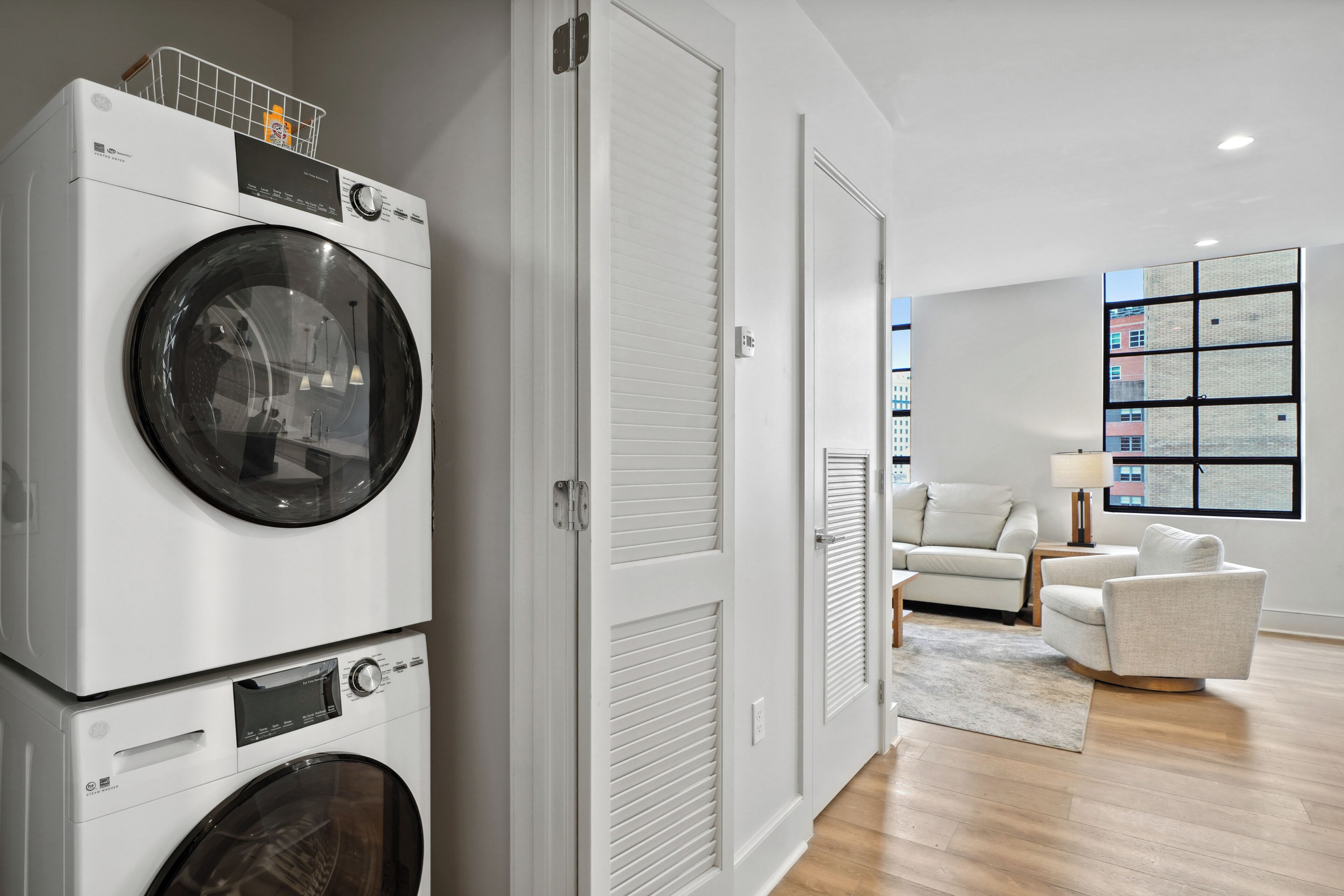 A laundry room with stacked washing machines, a closed door, and a living area with a sofa and a lamp.