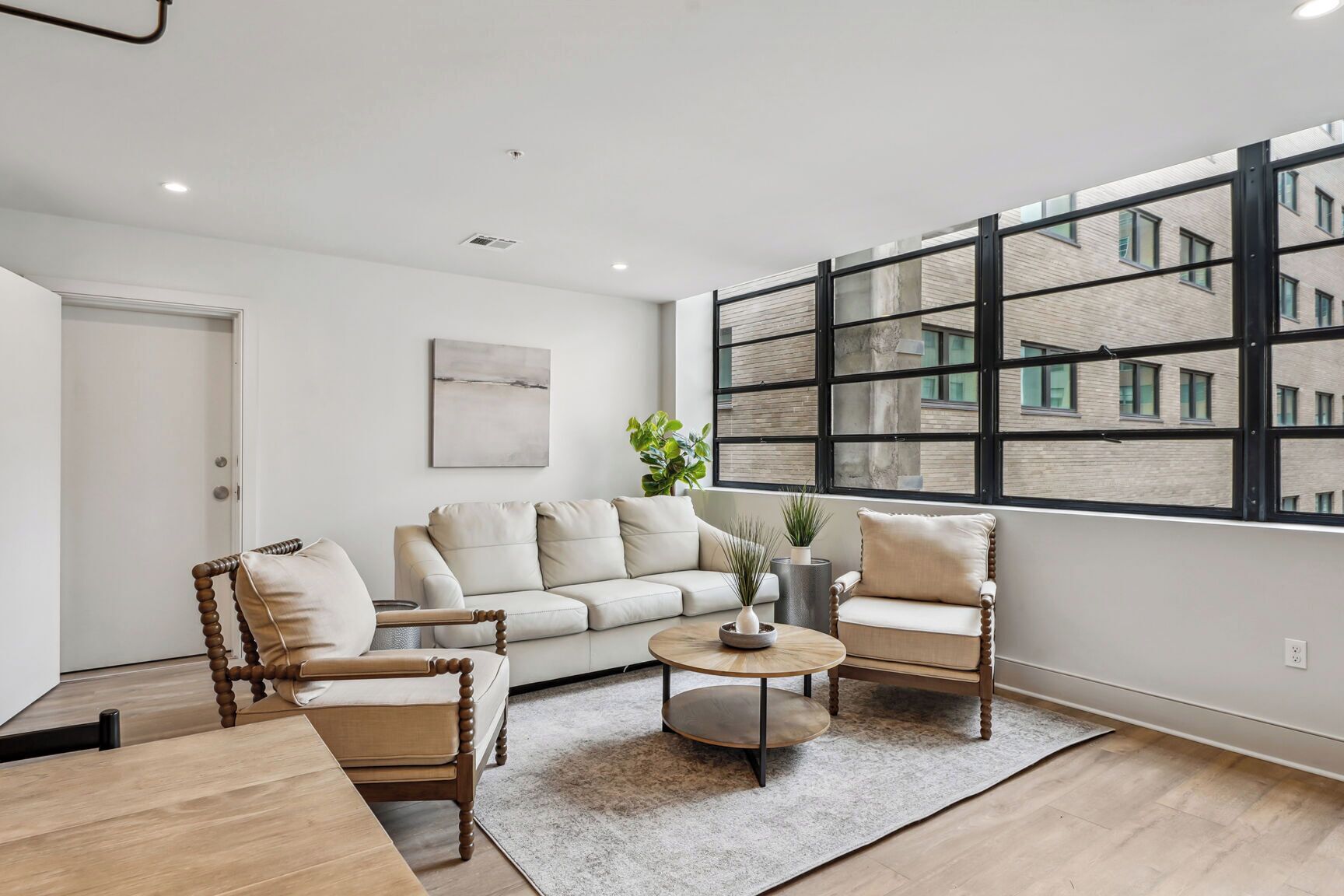A modern living room with a sofa, two armchairs, a coffee table, and a large window with a view of a brick building.