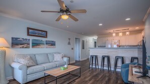 A modern living room with a sofa, coffee table, and a ceiling fan. A kitchen area with bar stools and a refrigerator is visible in the background.