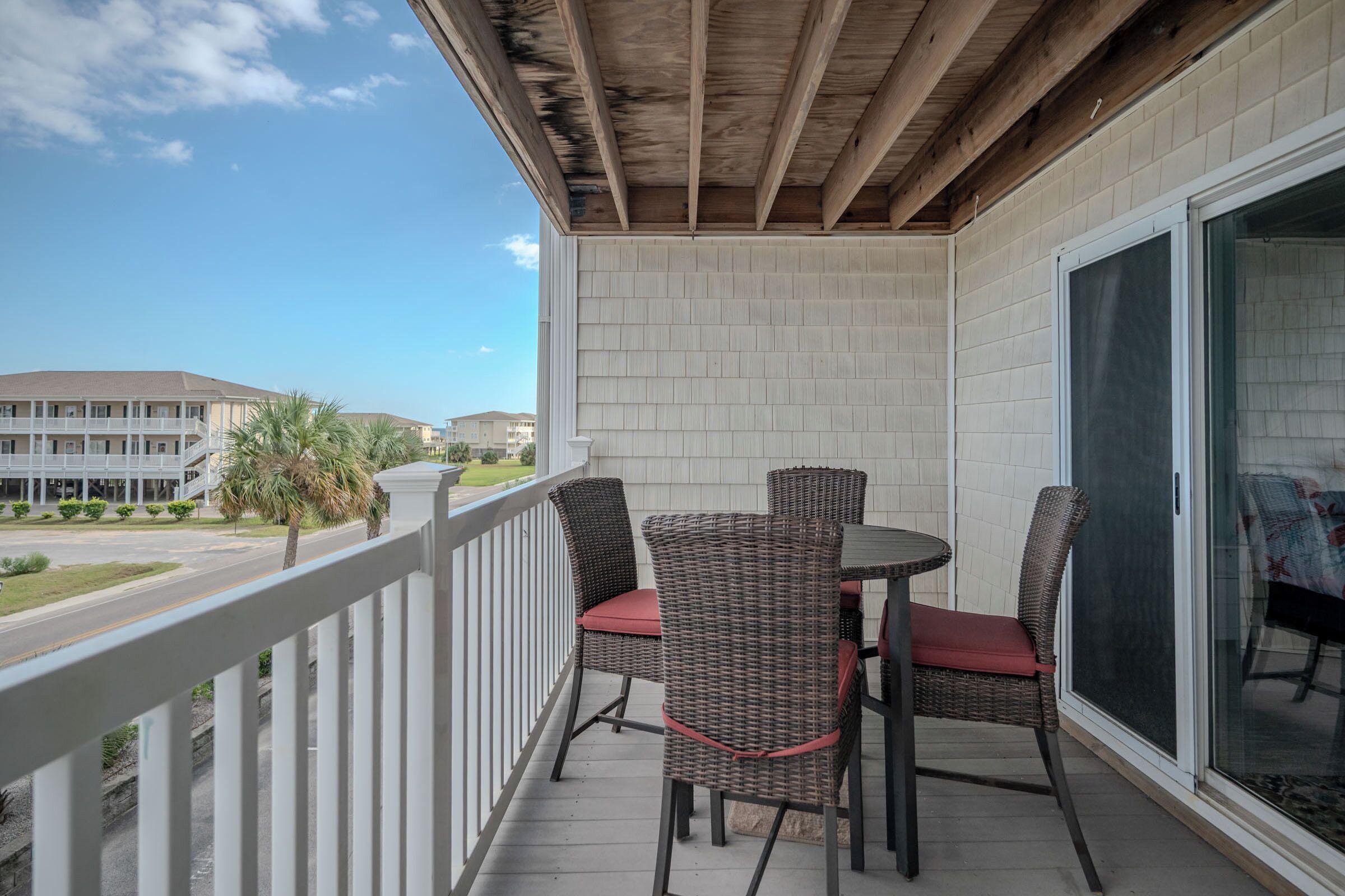 A balcony with wicker chairs and a table, overlooking a street with buildings and a palm tree.