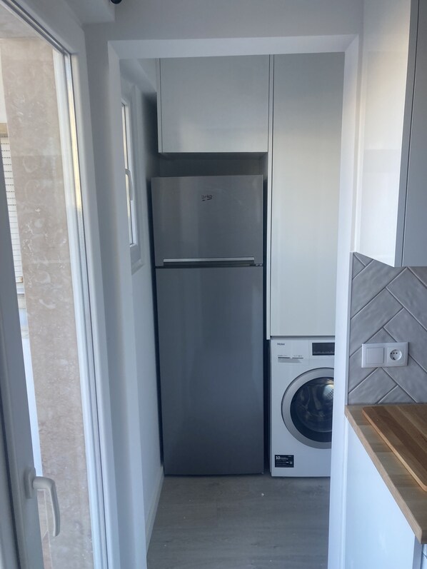 A compact kitchen with a stainless steel refrigerator, a washing machine, and a wooden countertop. - Apartamento com Terraço e 3 Quartos, 200 Metros da Praia (Costa da Caparica)