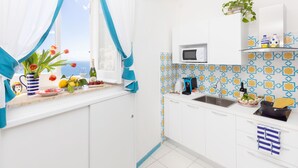 A bright kitchen with white cabinets, a window with blue curtains, a microwave, and a sink. - Adoro Positano (Positano)