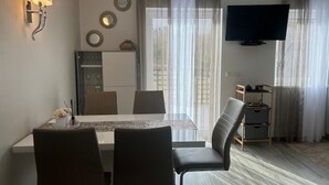 A modern living room with a dining area, a flat-screen TV mounted on the wall, and a large window with sheer curtains.