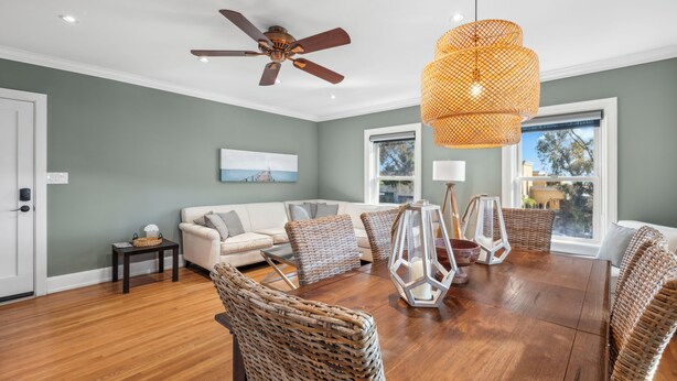 A spacious living room with a wooden floor, a ceiling fan, a large dining table, wicker chairs, and a hanging light fixture.