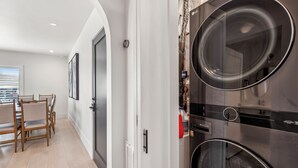 A laundry room with stacked washers and dryers, a door leading to a dining area with a table and chairs, and a window with blinds.