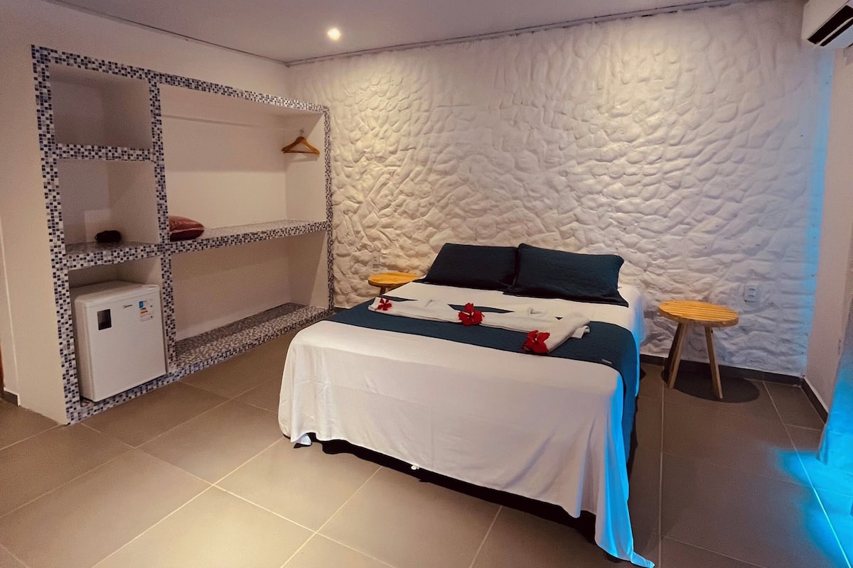 A bedroom with a bed, a bedside table, a hanging shelf, and a small stool.