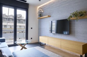 A modern living room with a large flat-screen TV mounted on a brick wall, a glass coffee table, and a wooden console table. - Acropolis Modern Apartment  (Athina)