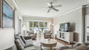 A spacious living room with a large gray sofa, a round coffee table, and a television on a cabinet. There is a ceiling fan, a floor lamp, and a view of trees through large windows.