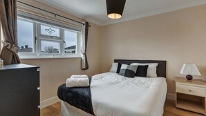 A bedroom with a bed, a nightstand, a dresser, a window with curtains, and a ceiling light. - 3-bed House, Sleeps 6, Free Parking,wifi (Basildon)