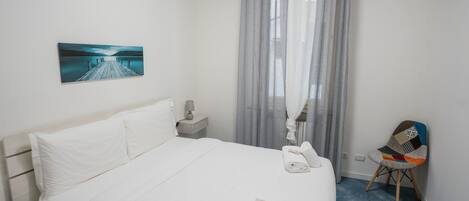 A hotel room with a bed, a nightstand, a chair, a painting, and a window with curtains.