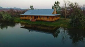 A wooden cabin with a large porch, situated by a calm lake with a reflection.