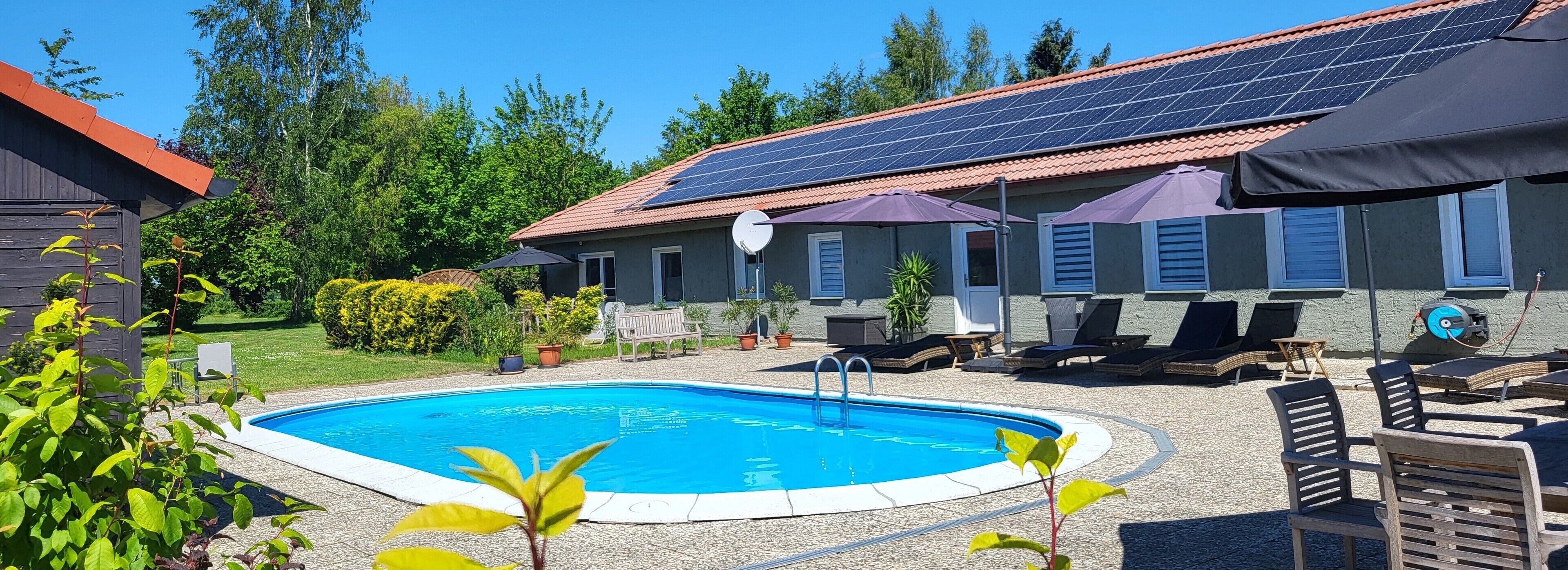 Una piscina con solárium de baldosas, rodeada de sillas de descanso y sombrillas, con un edificio que incluye paneles solares y techo de tejas al fondo.