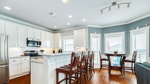 A kitchen with white cabinets, a central island with bar stools, and a dining area with a round table and chairs. - Sun, Sand & Surf Await! 3rd House from Beach, Dogs Welcome, Fenced Area, 2 Decks (Galveston)