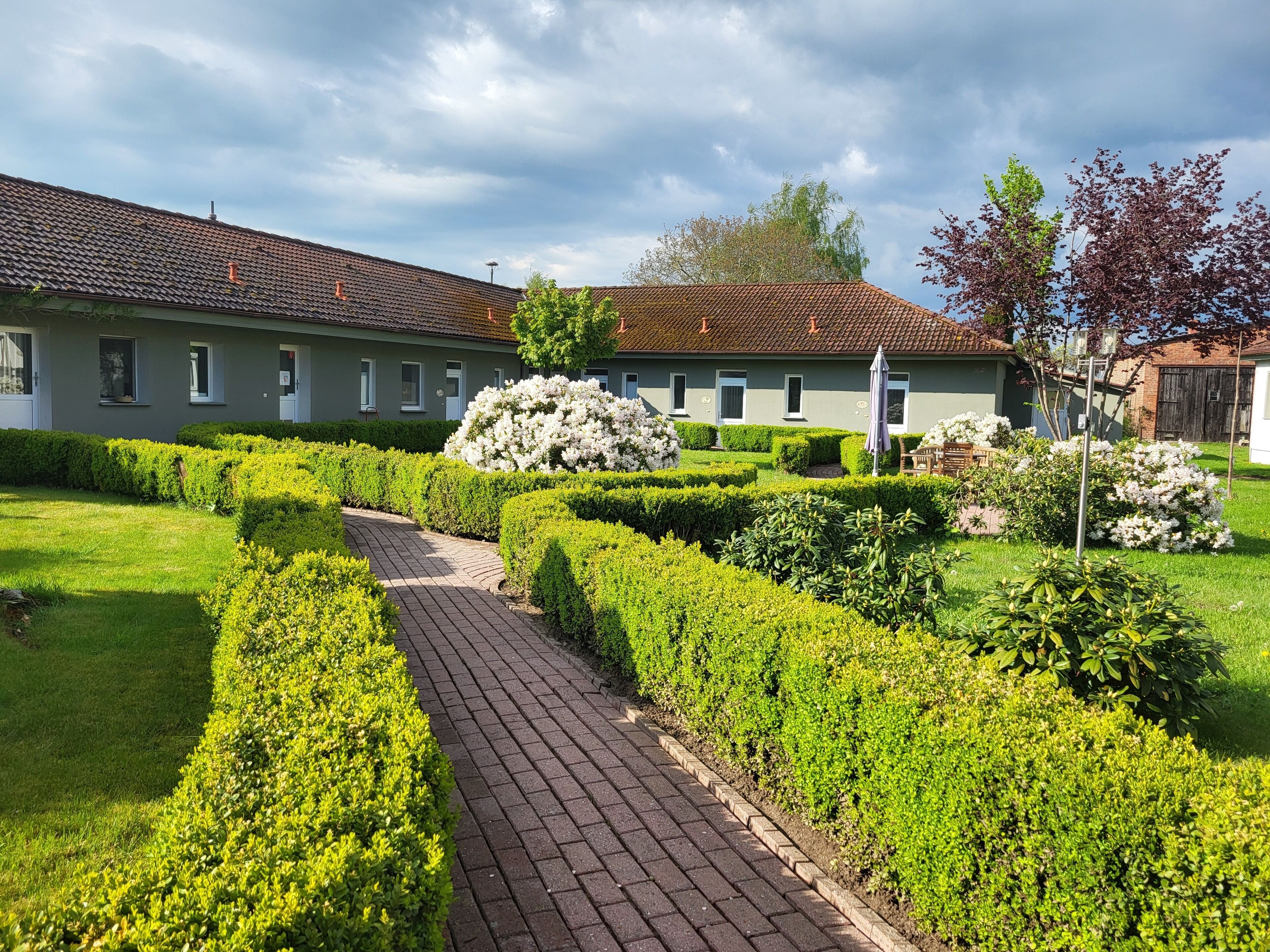 A courtyard with a paved pathway, neatly trimmed hedges, and a building with a tiled roof.