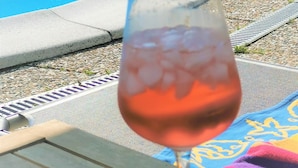 Un vaso de bebida rosada en una silla de descanso junto a una piscina con borde azul.