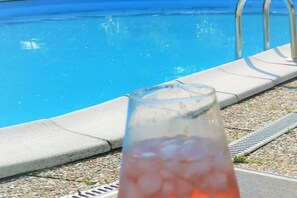 Un verre d’une boisson rouge, avec des glaçons, est posé à côté d’une piscine dont l’eau est d’un bleu limpide, et on aperçoit une échelle de piscine en métal.