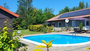 Een zwembad met betegelde rand, omringd door ligbedden en parasols, met op de achtergrond een gebouw met zonnepanelen en een betegeld dak.