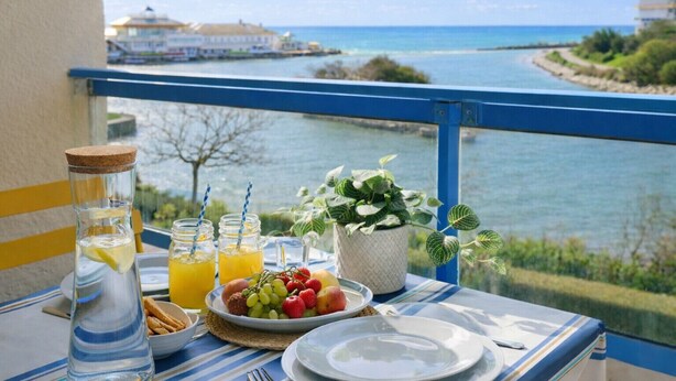 A balcony table with a view of a river, a bottle of water, a glass of orange juice, a plate of fruit, and a small potted plant. - Pretty flat with ocean and lake views Les Sables - sleeps 4 (Les Sables-d´Olonne)