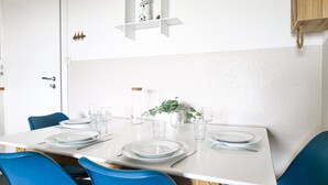 A dining table set with plates, glasses, and cutlery, surrounded by blue chairs, with a plant on the table and a white wall with a shelf above. - Pretty flat with ocean and lake views Les Sables - sleeps 4 (Les Sables-d´Olonne)