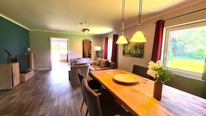 A spacious living area with a wooden dining table, comfortable seating, and a large window offering a view of greenery.