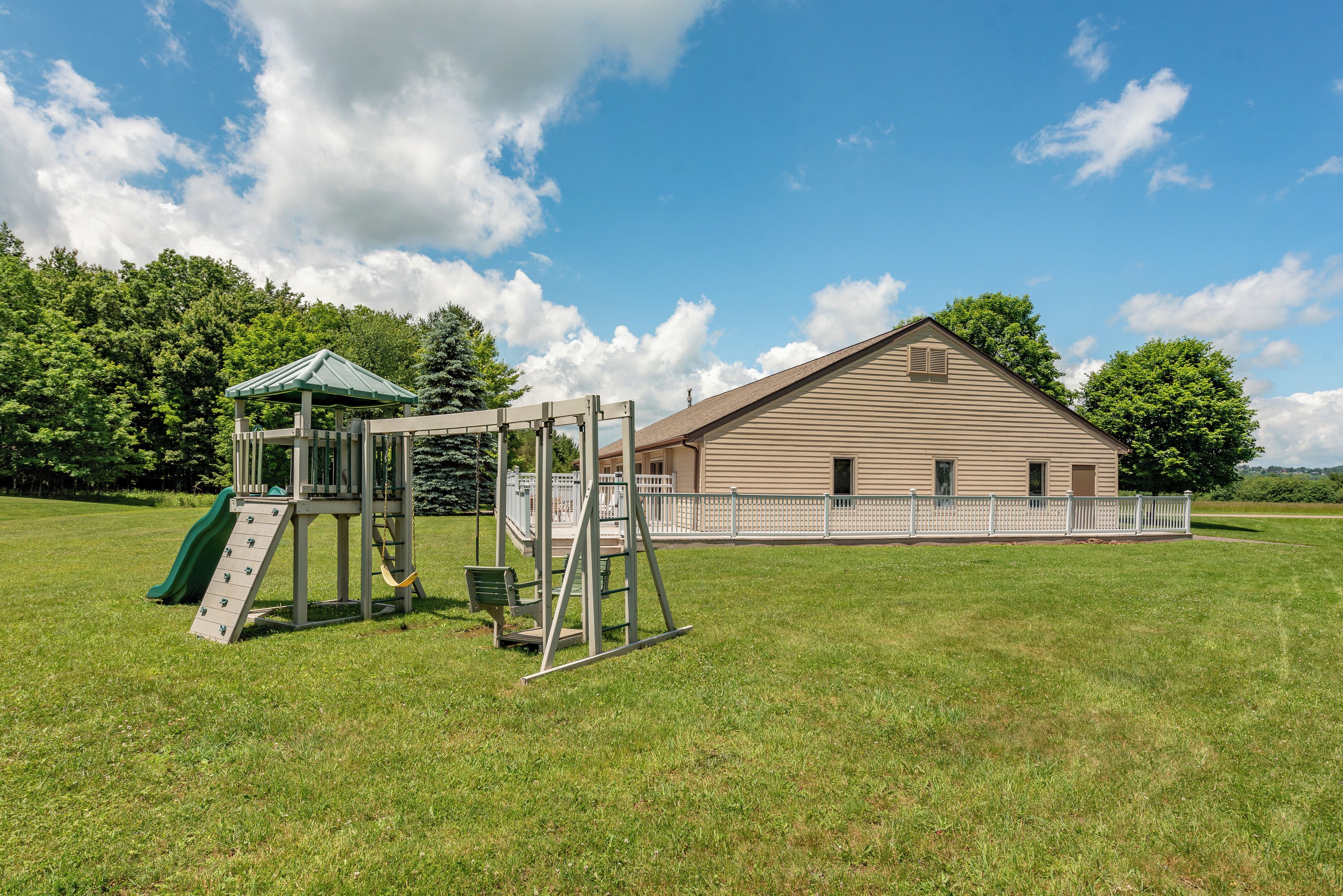 A playground with climbing structures and a slide, a white fence, and a large building in the background.