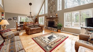 Un salon spacieux doté d’une cheminée en pierre, de parquets en bois et de grandes fenêtres offrant une vue sur l’extérieur.