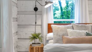A bedroom with a wooden headboard, white bedding, a bedside lamp, a small wooden nightstand with a plant, and a window with a view of trees. - Luxury Modern Cabin* 5 Acres* Pickle Ball*Hot Tub (Primm Springs)