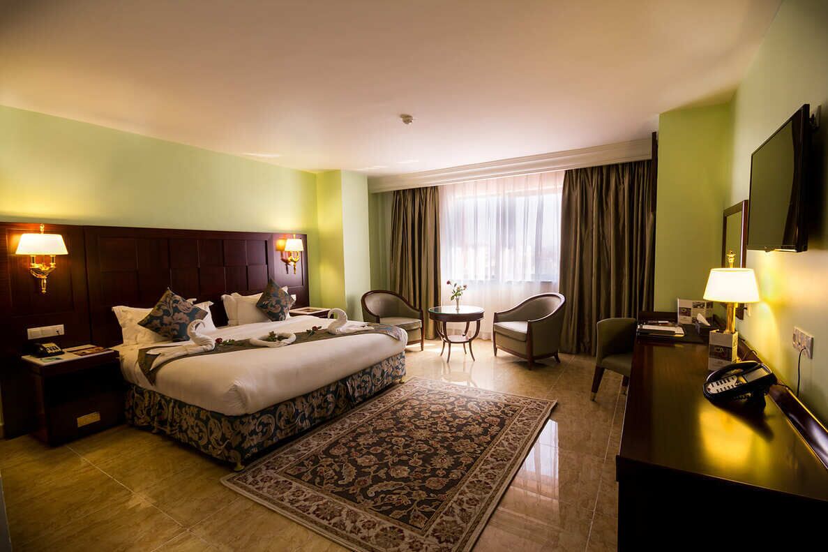 Habitación de hotel con una cama grande, mesitas de noche, un escritorio, una silla, un televisor y una ventana con cortinas.