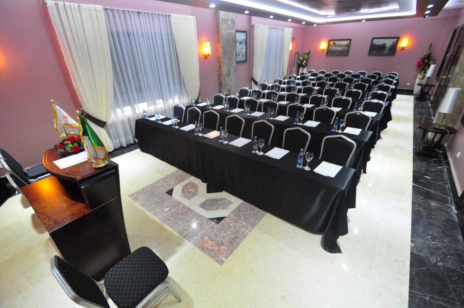Un salón de conferencias con filas de sillas, una mesa larga con tarjetas de nombre y un atril con micrófono.