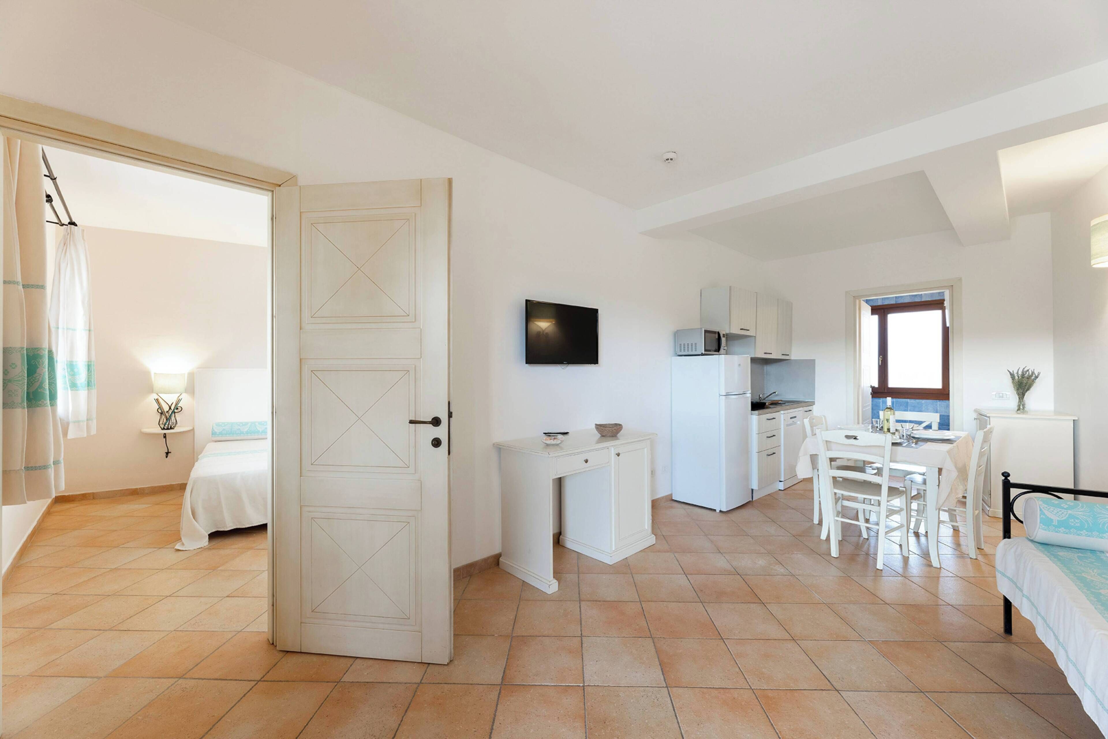 A room with a bed, a desk, a dining table with chairs, a TV, and a kitchenette.
