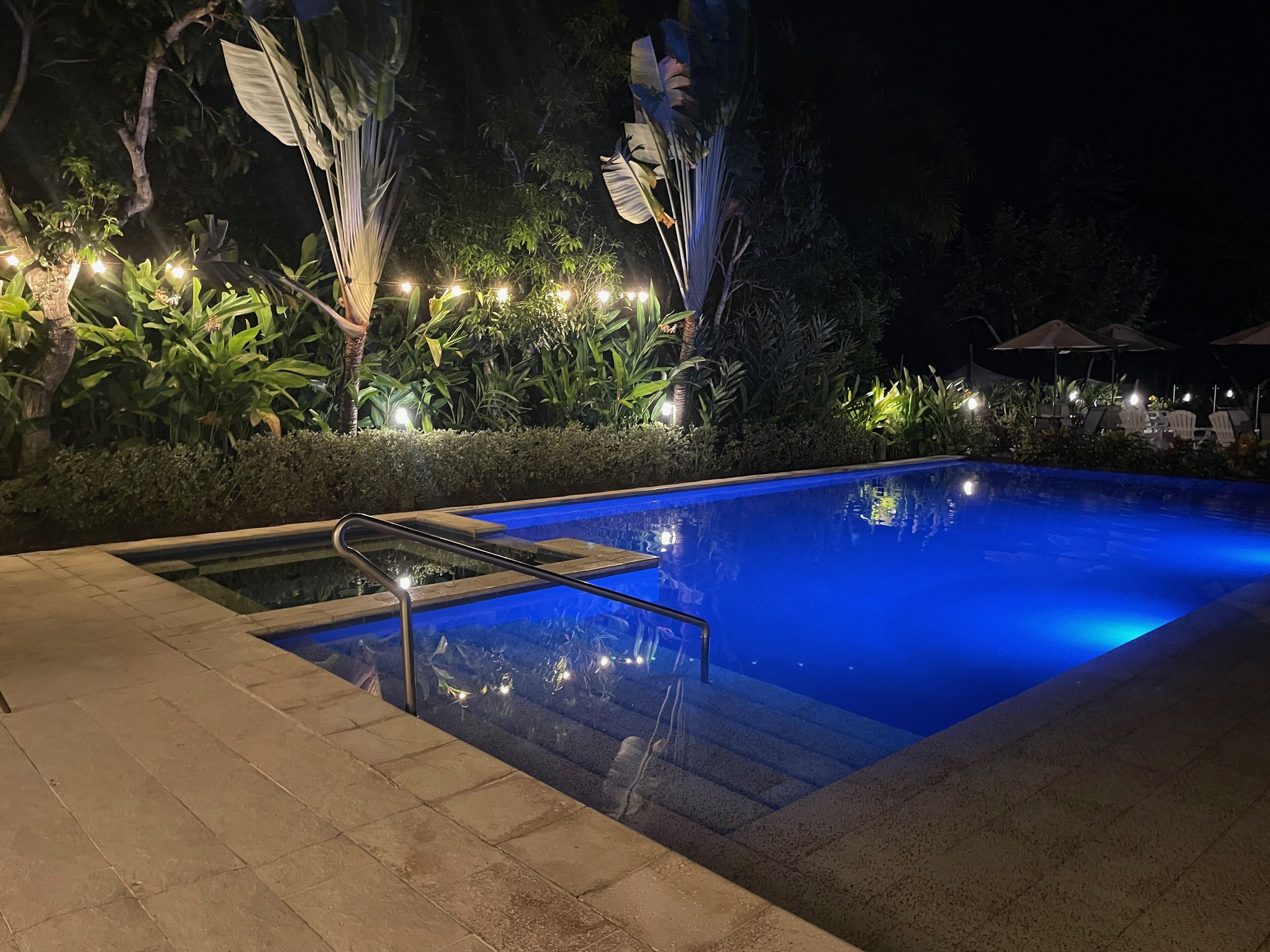 A nighttime swimming pool area with a clear pool, steps leading into the water, and surrounding greenery.