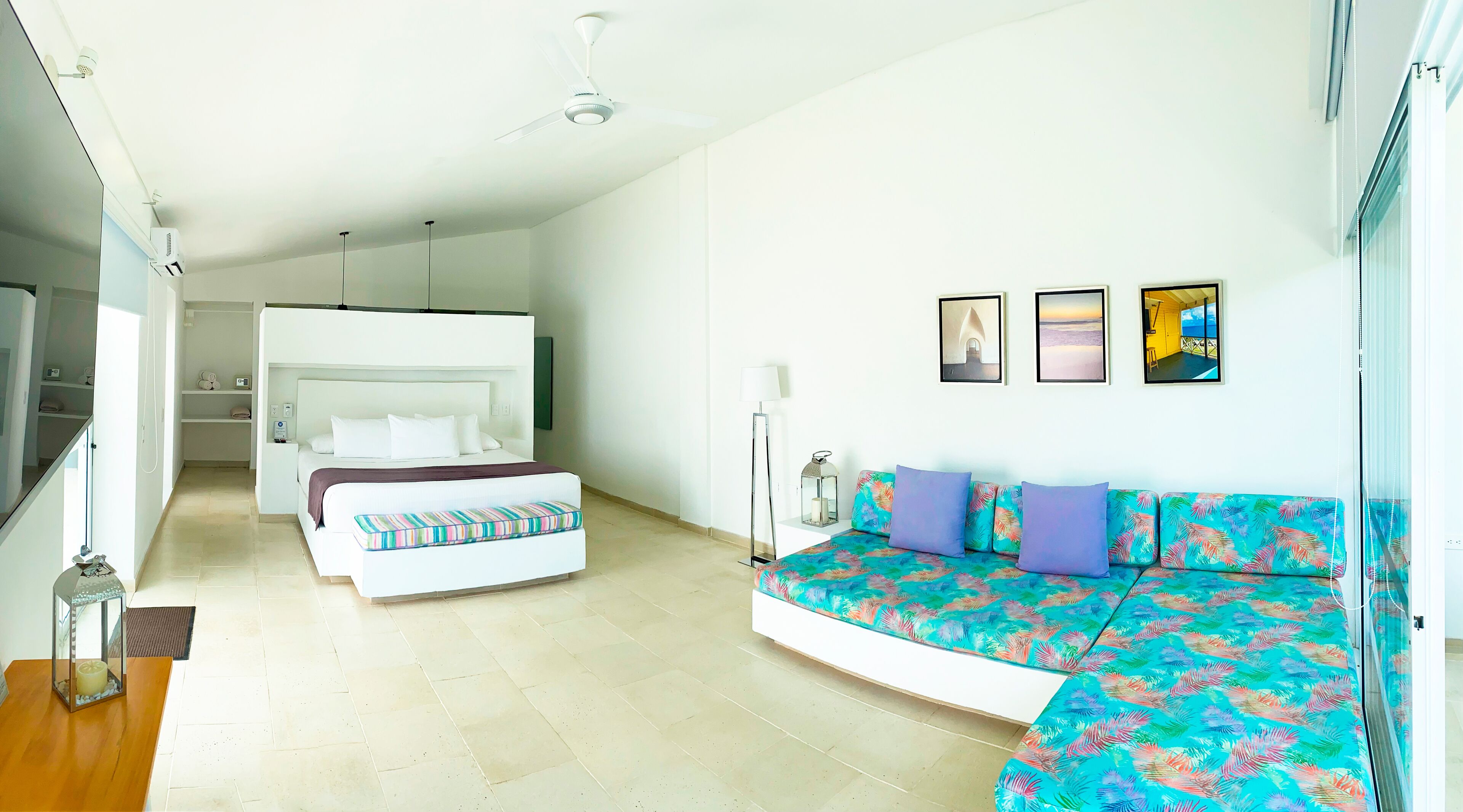 A modern bedroom with a bed, a sofa, and a nightstand. There are ceiling fans, a floor lamp, and a window with blinds.