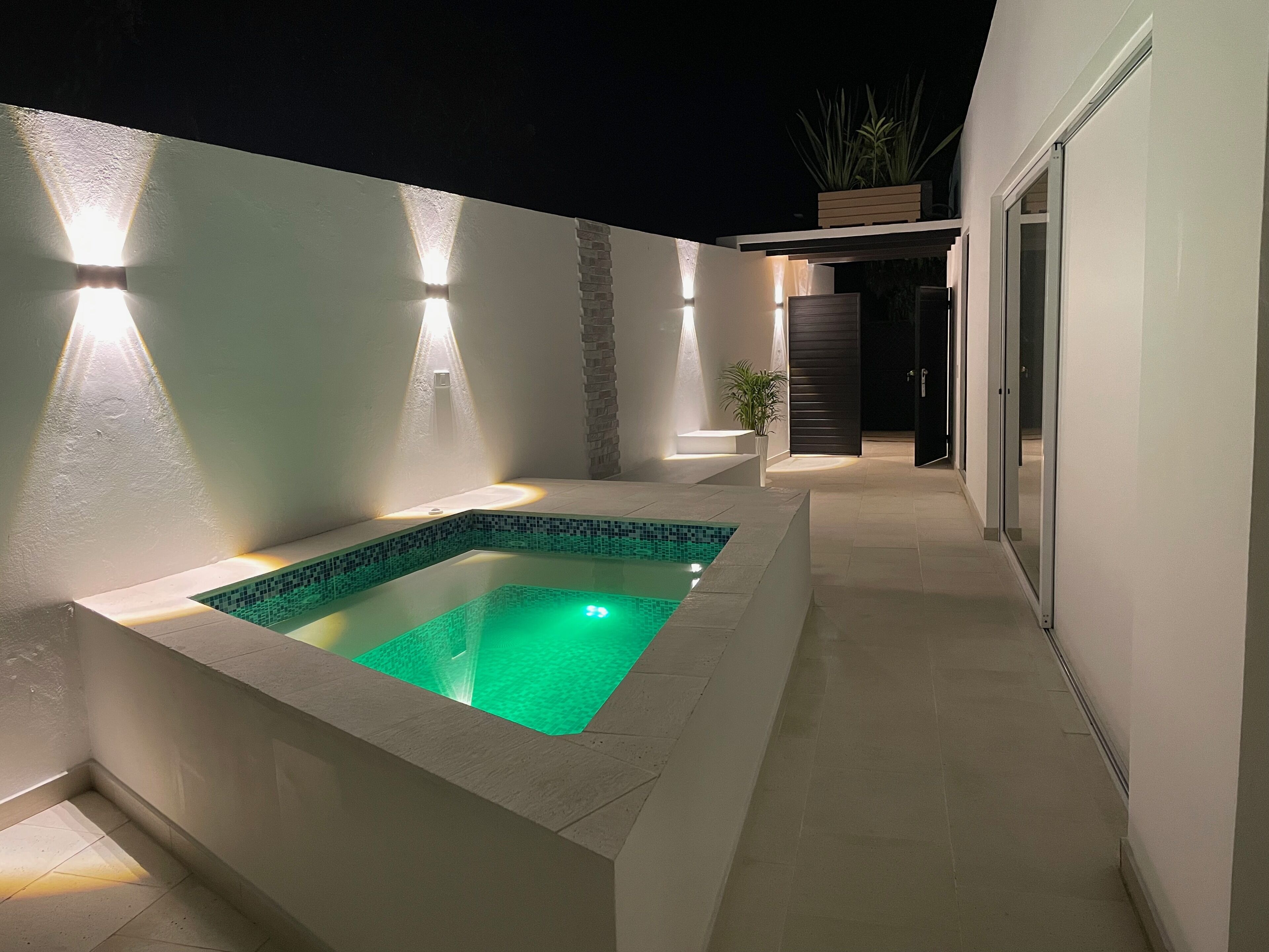 Un área de piscina exterior moderna con una piscina rectangular, rodeada por un muro blanco y un muro de acento de piedra. Hay luces que iluminan el espacio, y una puerta que da a otra parte de la propiedad.