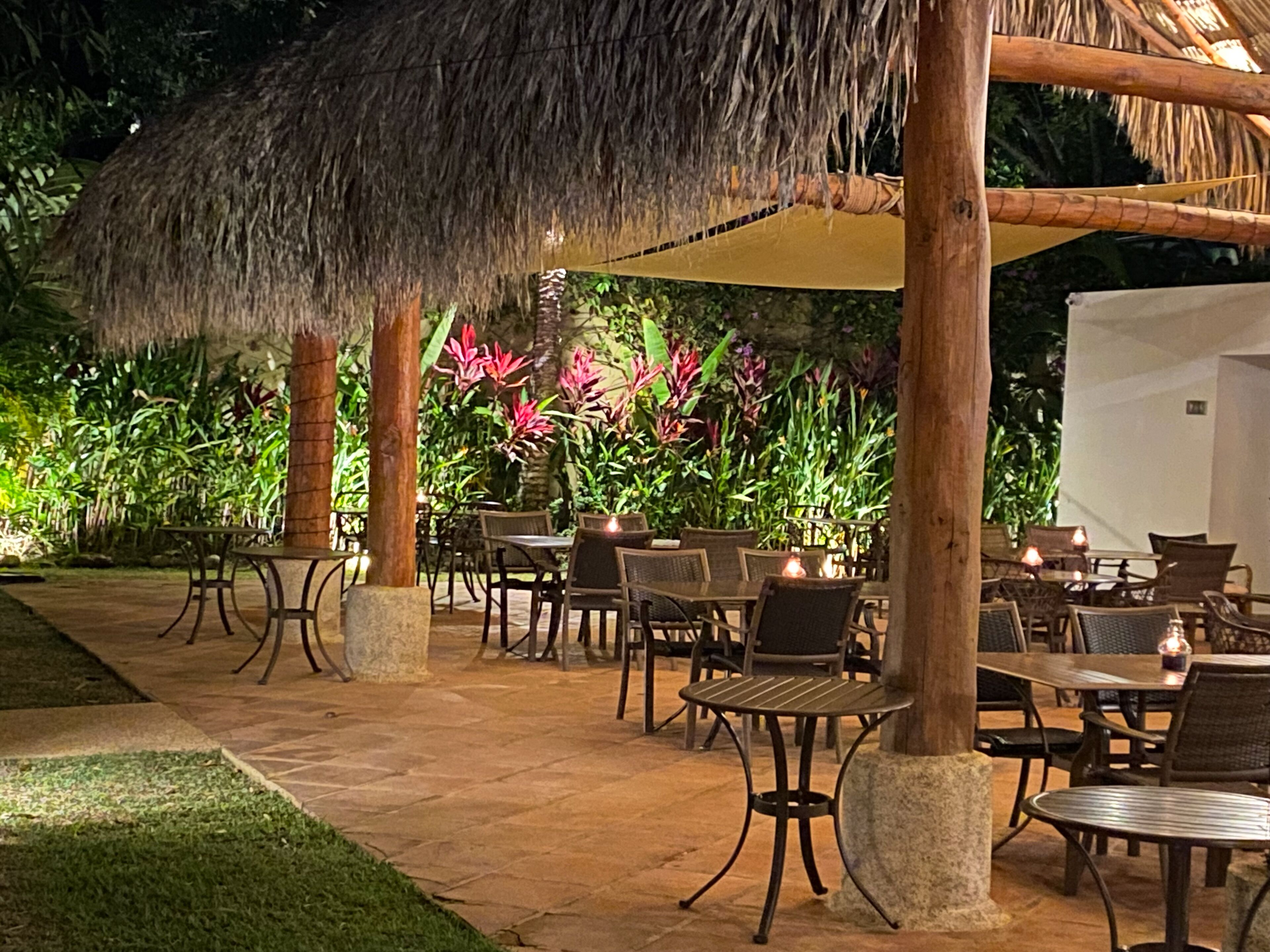 Un patio al aire libre con mesas y sillas, rodeado de abundante vegetación y una estructura de techo de paja.