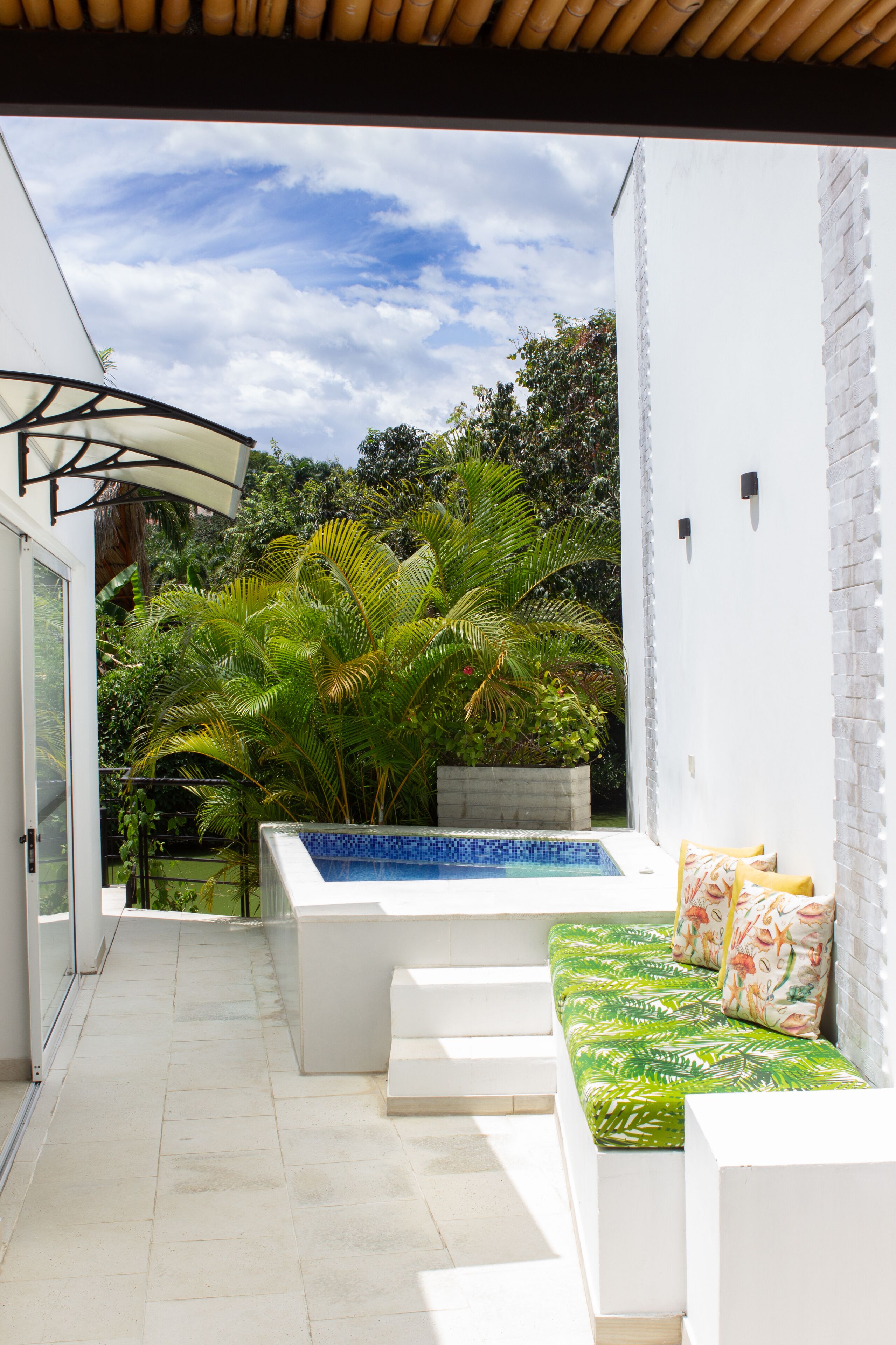A modern outdoor patio with a white wall, a built-in bench with green cushions, and a small pool.