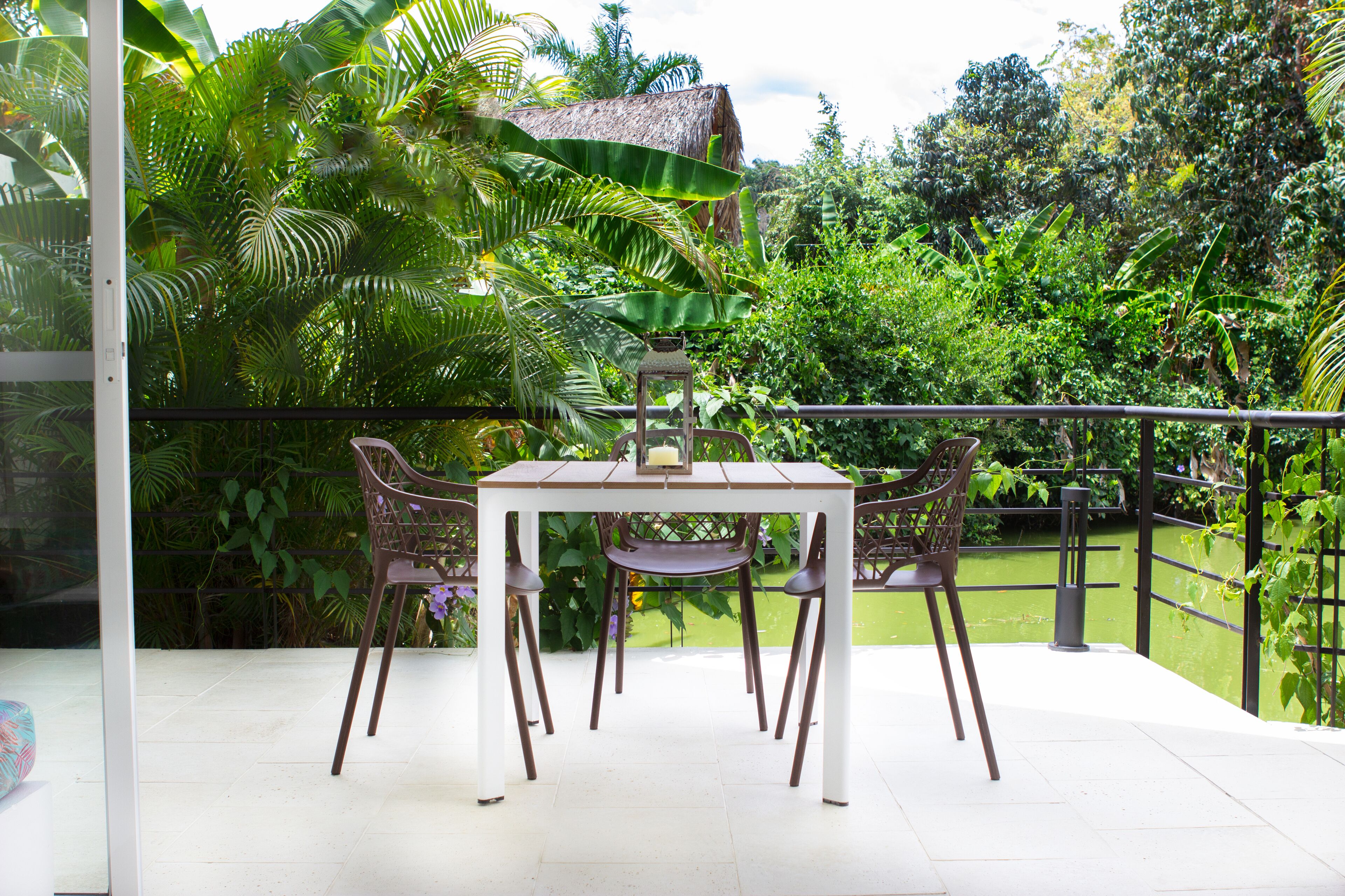 A balcony with a white table and four chairs, surrounded by lush greenery and a thatched roof structure.