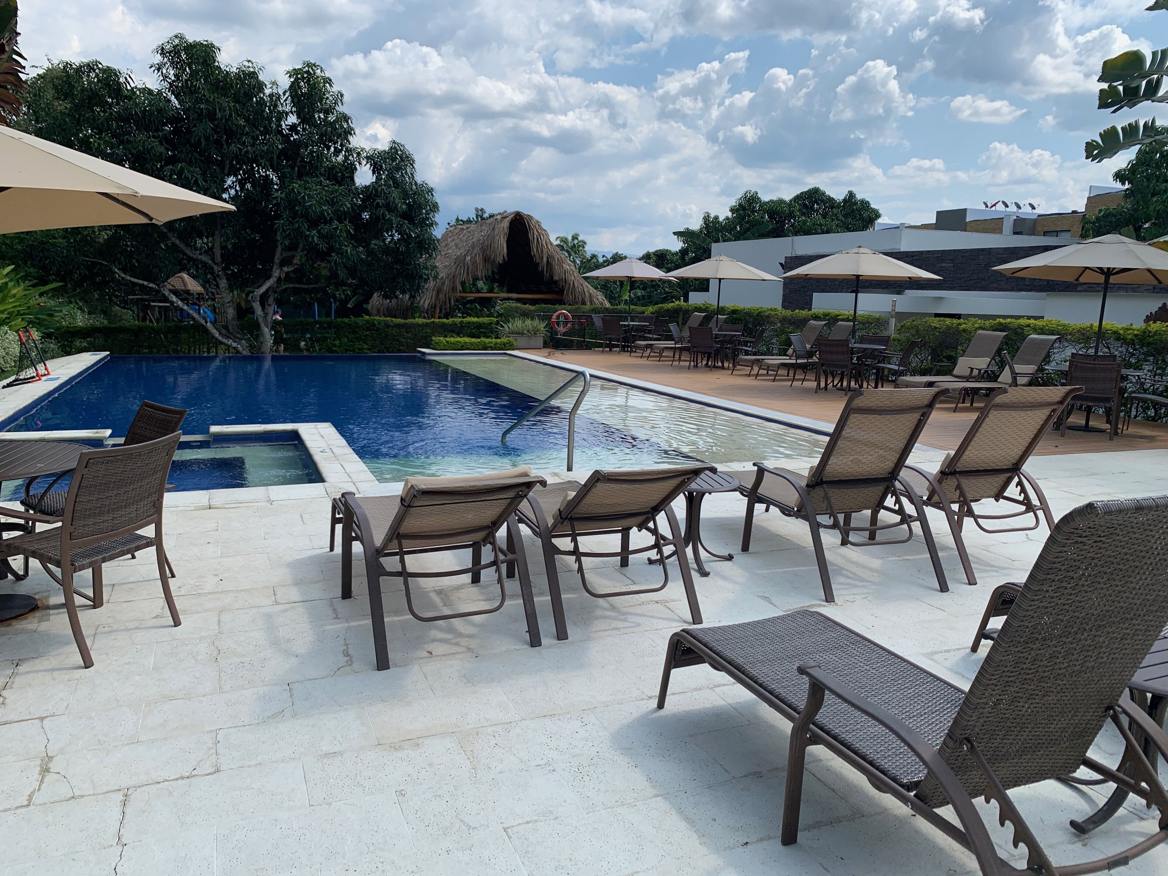 A poolside area with lounge chairs and umbrellas, surrounded by greenery and a building in the background.