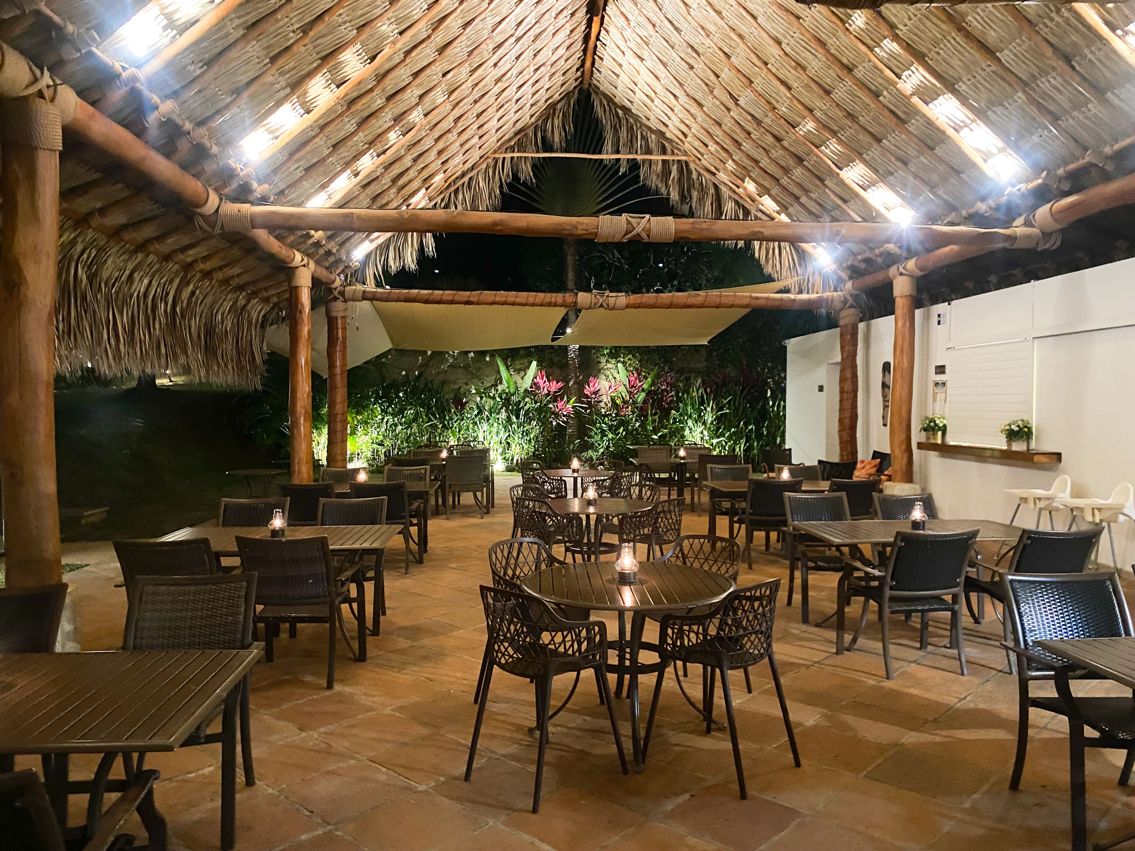 Un área de comedor al aire libre con techo de paja, mobiliario de mimbre y suelo de piedra.