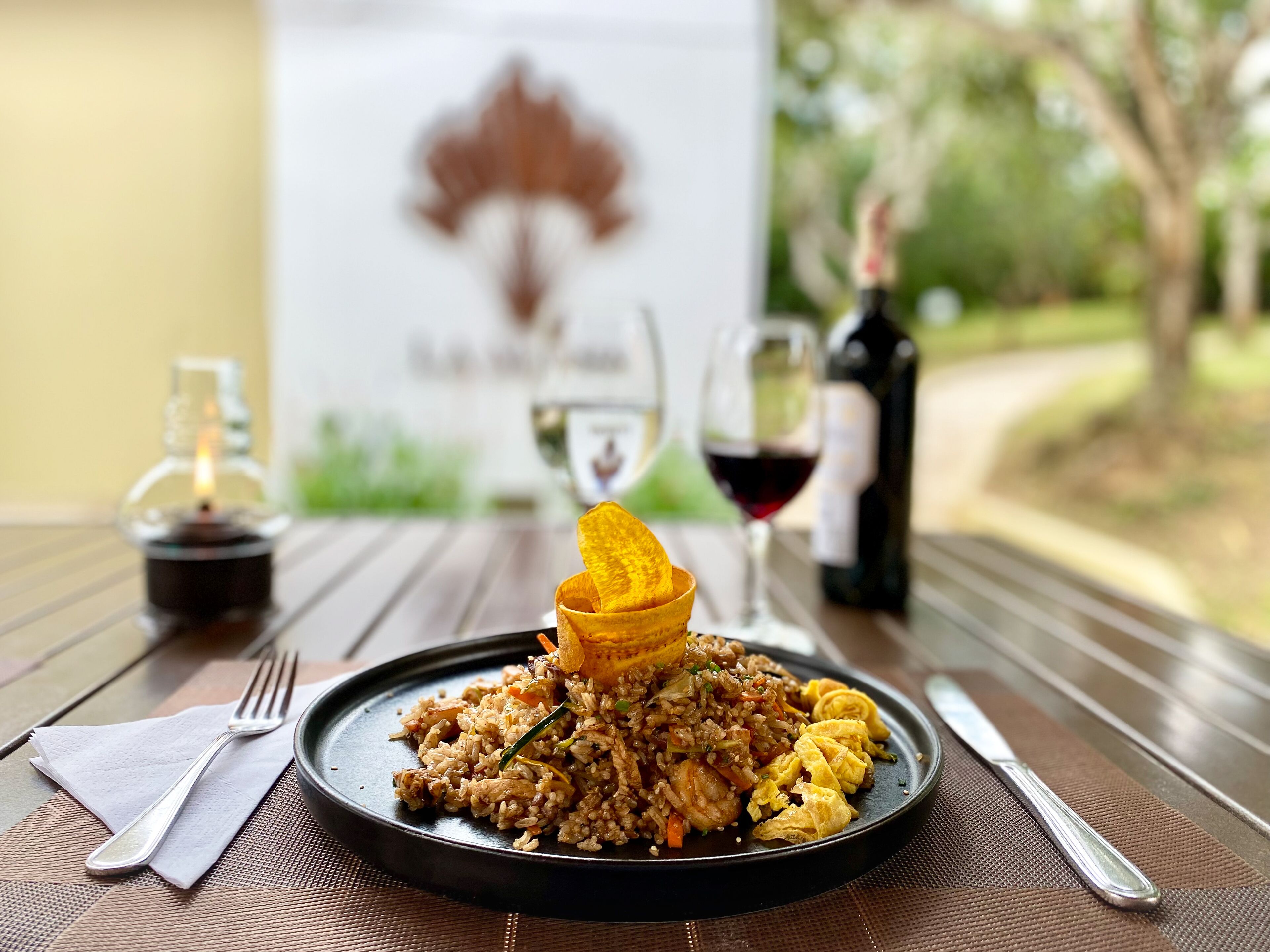 A plate of fried rice with vegetables and scrambled eggs on a black plate, accompanied by a bottle of wine and a glass of red wine on a wooden table.