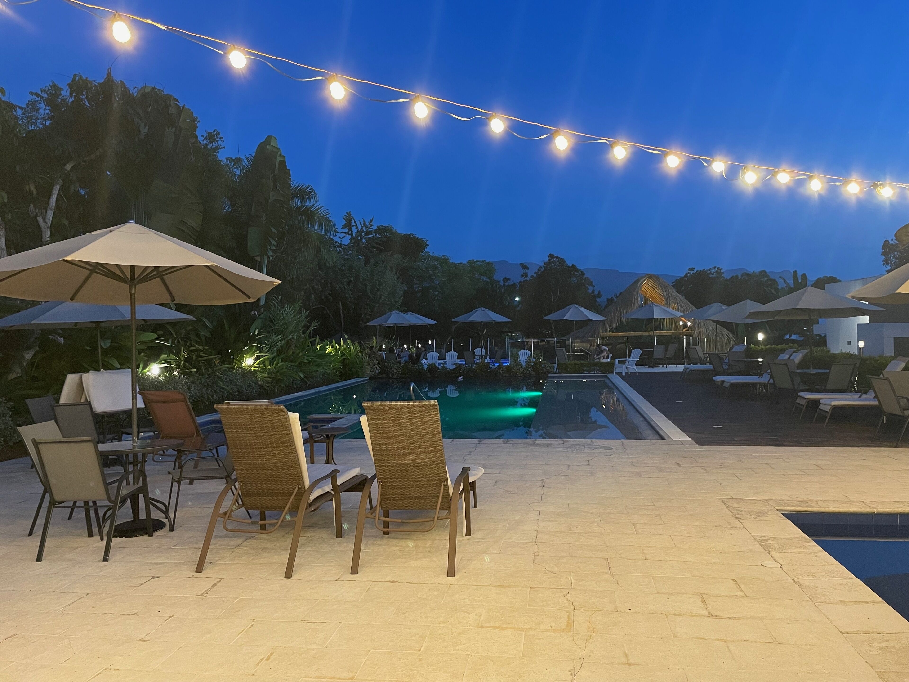 An outdoor pool area with lounge chairs and umbrellas, string lights overhead, and a view of the pool.