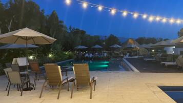 An outdoor pool area with lounge chairs and umbrellas, string lights overhead, and a view of the pool.