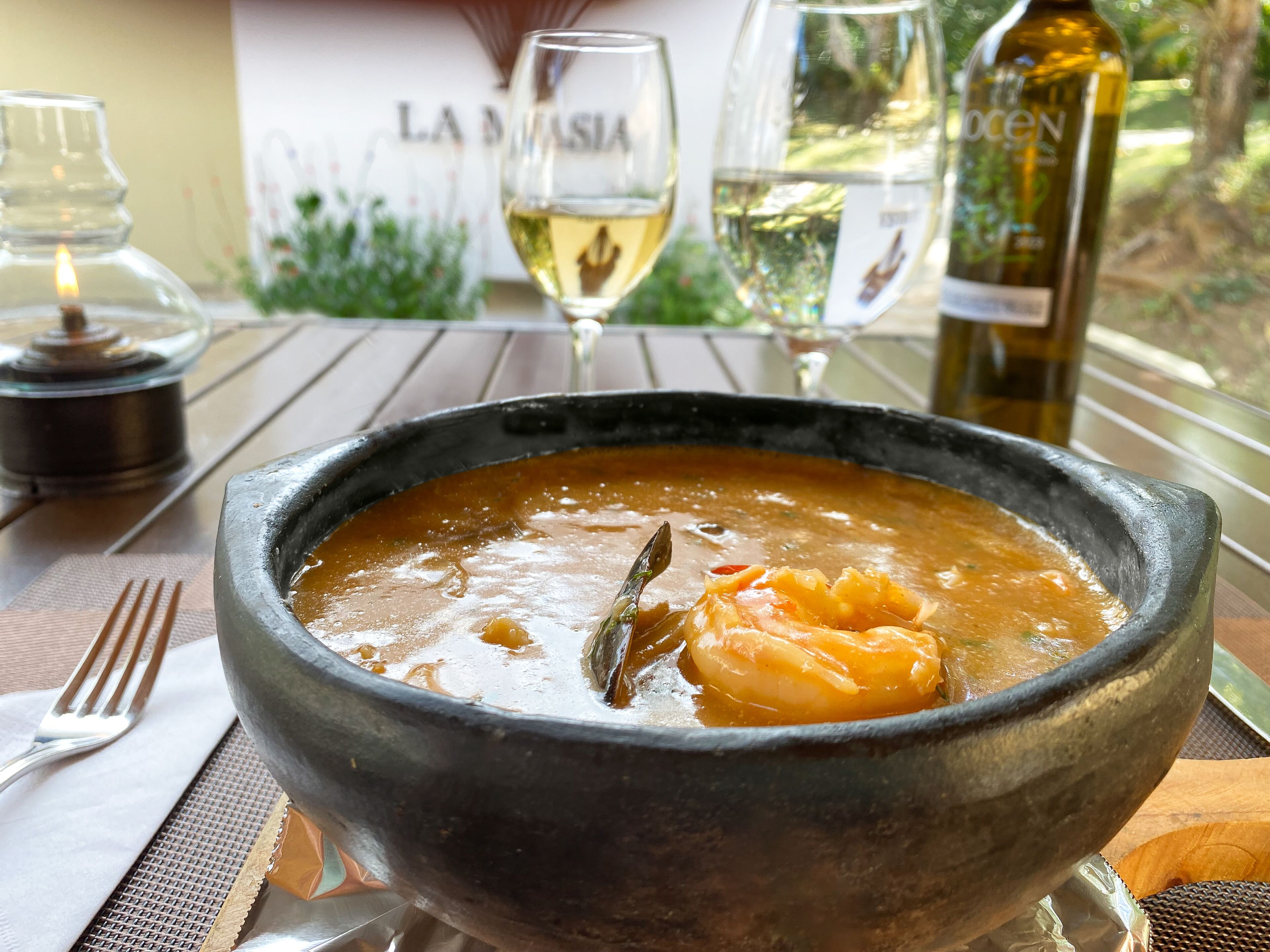 A bowl of soup with a bay leaf on a table with a fork, a candle, and a wine bottle.